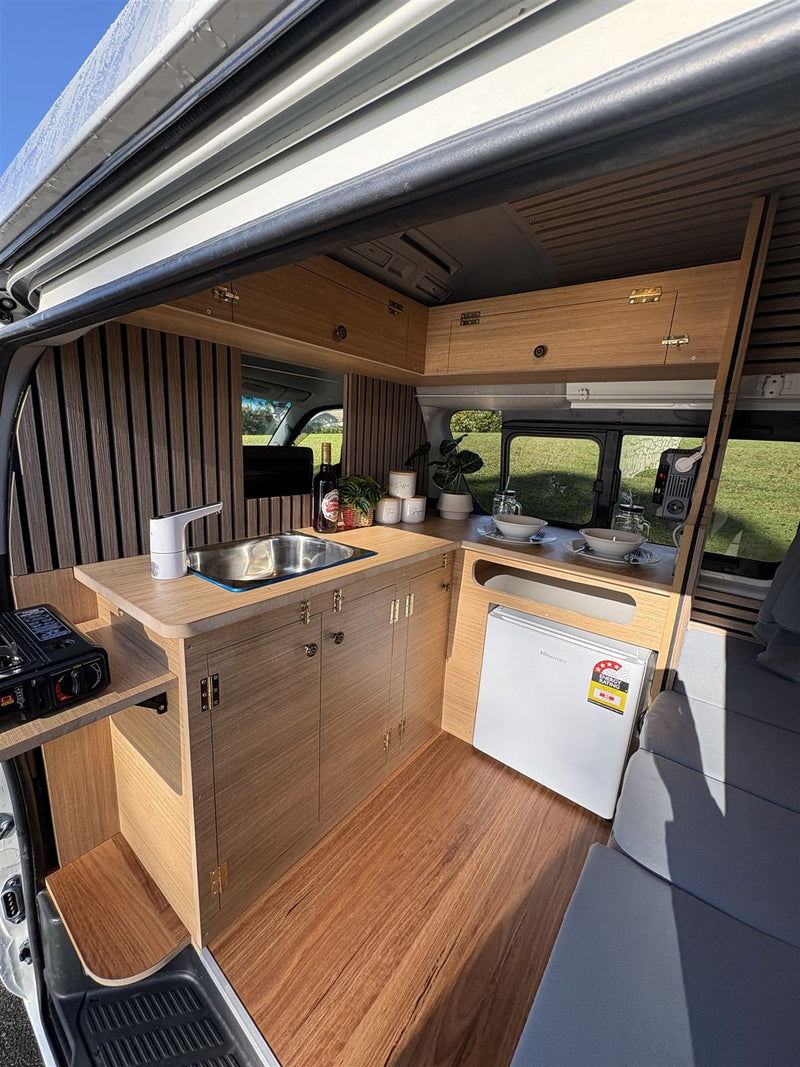 Campervan kitchen with sink, running water, fridge and timber cabinetry, seen through the open side door