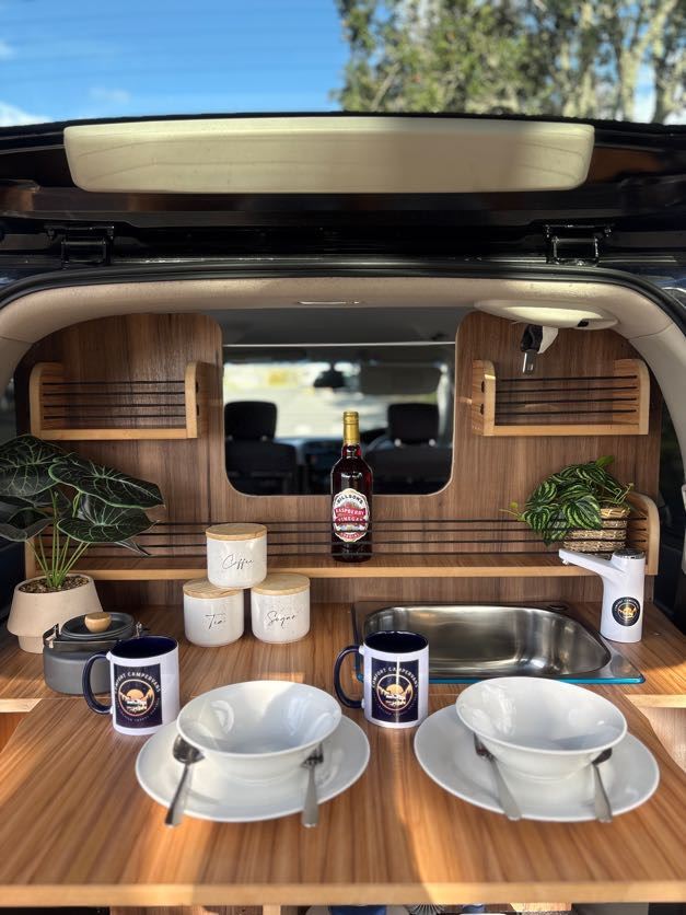 Rear kitchen of a campervan set for a meal with bowls, mugs and canisters under the open tailgate