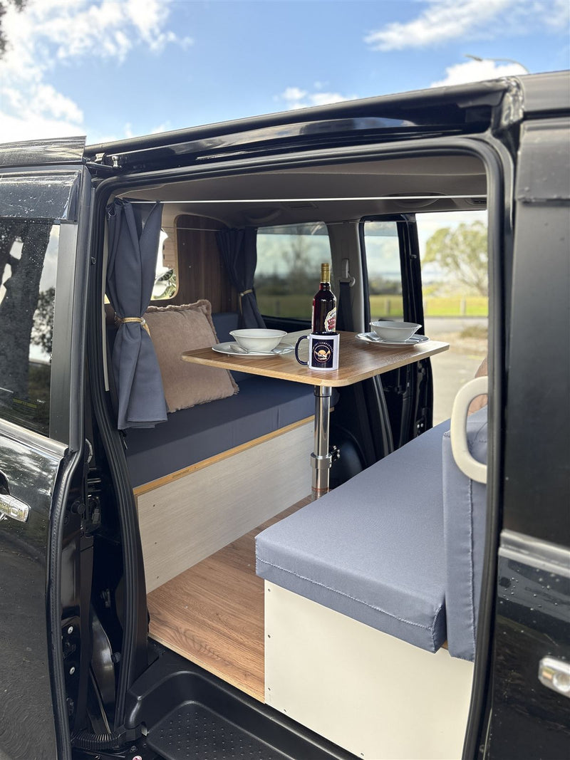 Side door of a black campervan open to a made bed, table set for two and a view of open sky