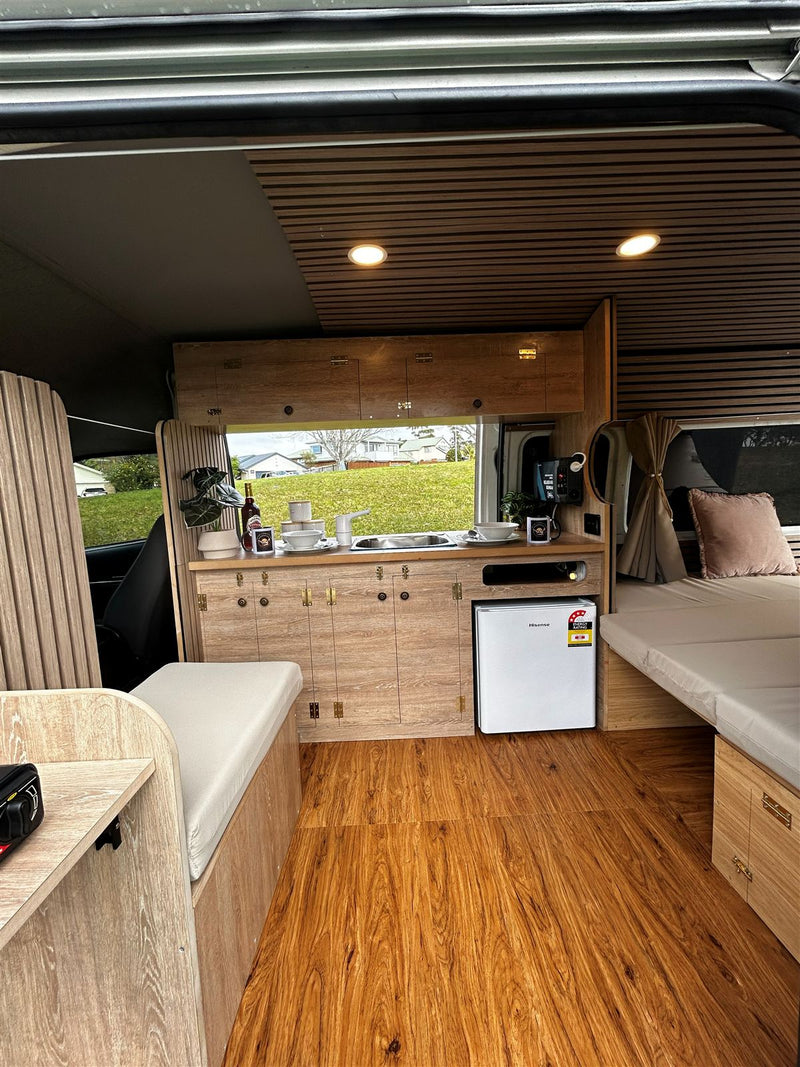 Campervan kitchen bench with sink and fridge, seating on one side and the bed on the other